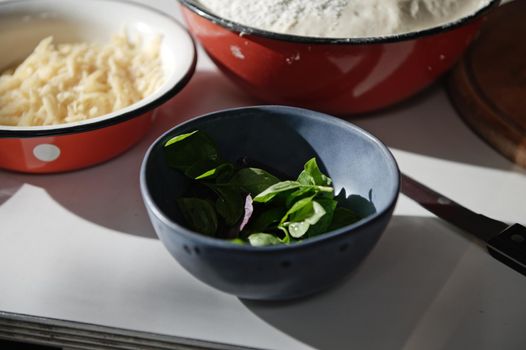 Close-up. Freshly picked basil leaves - fragrant culinary herbs in a navy blue ceramic bowl, nearby vintage bowls with grated cheese and raising yeast dough on white table background. Food still life