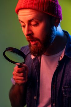 Hipster guy looking through magnifying glass, spying, finding out something, exploring, inspecting, wearing beanie hat and denim vest. Indoor studio shot isolated on colorful neon light background.