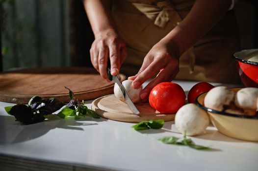 Details: Chef's hands using a kitchen knife, slicing fresh mushroom champignon on a chopping board. Fragrant basil leaves lying on a kitchen countertop in a rustic kitchen . Chopped pizza ingredients.