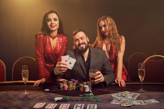 Two sexy women and handsome man are playing poker at casino. They are showing the winning combination, smiling and looking at the camera while posing at the table against a yellow backlight on smoke background. Cards, chips, money, gambling, entertainment concept.