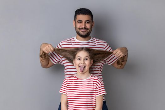 Portrait of happy funny father and daughter having fun, cute kid showing tongue out, dad pulling her hair, expressing positive emotions. Indoor studio shot isolated on gray background.