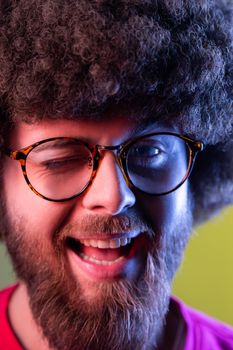 Closeup portrait of positive hipster man with Afro hairstyle in good mood, smiling broadly and winking at camera, keeps mouth open. Indoor studio shot isolated on colorful neon light background.
