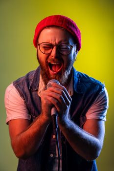 Happy positive hipster man with beard standing with microphone in both hands, singing songs, wearing beanie hat and denim vest. Indoor studio shot isolated on colorful neon light background.