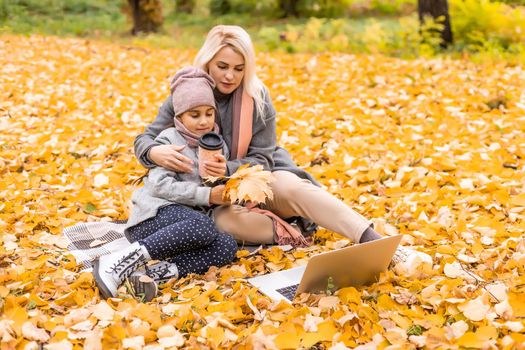 Mom and daughter resting on nature with laptop sitting on an autumn day.
