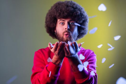 Portrait of hipster man with Afro hairstyle blowing heart shaped confetti, enjoying birthday or valentines day, wearing red sweatshirt. Indoor studio shot isolated on colorful neon light background.