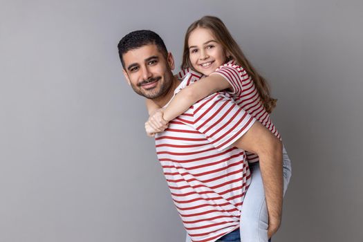 Portrait of smiling father and cute child in striped T-shirts posing together, dad give piggyback daughter, holding on back adorable kid. Indoor studio shot isolated on gray background.