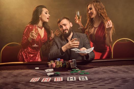 Two attractive females and good-looking male are playing poker at casino. They are holding their cash winning, smiling and having a good time while posing at the table against a yellow backlight on smoke background. Cards, chips, money, gambling, entertainment concept.