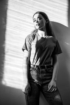Young adult beautiful woman wearing T-shirt and jeans standing looking at camera with dreamy romantic expression. Black and white photography, indoor studio shot illuminated by sunlight from window