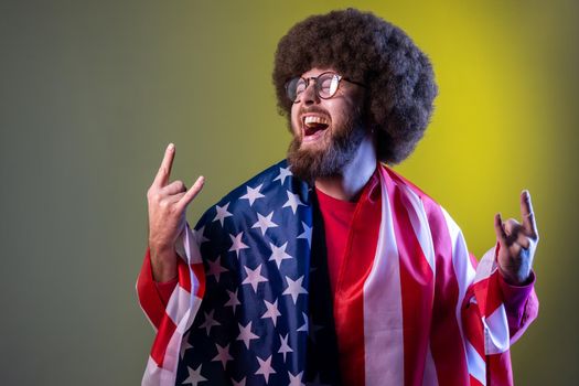 Excited happy hipster man with Afro hairstyle wrapped in american flag, rejoicing, celebrating national holiday, showing v sign. Indoor studio shot isolated on colorful neon light background.