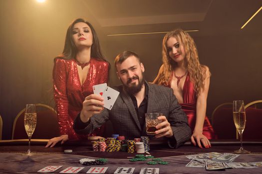 Two sexy females and handsome male are playing poker at casino. They are showing the aces, smiling and looking at the camera while posing at the table against a yellow backlight on smoke background. Cards, chips, money, gambling, entertainment concept.