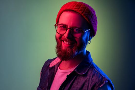 Extremely happy bearded hipster man looking at camera with toothy smile, optimism, success, wearing red beanie hat and denim vest. Indoor studio shot isolated on colorful neon light background.