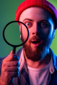 Portrait of amazed hipster man standing, holding magnifying glass and looking at camera, having astonished surprised face. Indoor studio shot isolated on colorful neon light background.