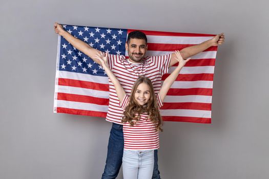 Satisfied father and daughter standing with raised arms, man holding big american flag, family celebrating national holiday or relocation in USA. Indoor studio shot isolated on gray background.