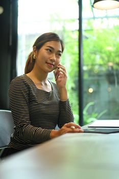 Charming asian female freelancer sitting in coffee shop and talking on mobile phone.