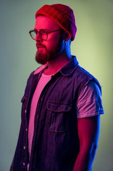 Portrait of confident bearded male looking to side space with serious attentive face, wearing beanie hat and denim vest. Indoor studio shot isolated on colorful neon light background.