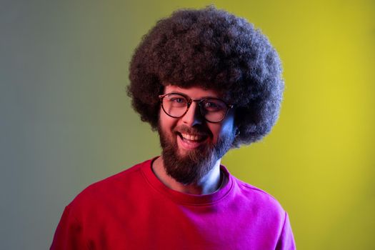 Portrait of hipster man with Afro hairstyle looking at camera with happy smile, has good mood, wearing red sweatshirt. Indoor studio shot isolated on colorful neon light background.