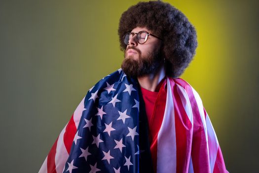 Portrait of proud hipster man with Afro hairstyle standing wrapped in american flag, looking away, feels pride of his USA. Indoor studio shot isolated on colorful neon light background.