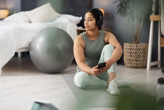 Taking breaks with her connections. a sporty young woman wearing headphones and using a cellphone while exercising at home