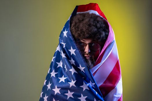 Portrait of hipster man with Afro hairstyle standing wrapped in american flag, looking away with funny facial expression and pout lips. Indoor studio shot isolated on colorful neon light background.