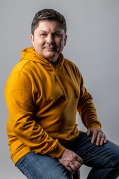 Portrait of handsome optimistic middle aged man looking at camera, sitting on chair and expressing positive emotions, wearing urban style hoodie. Indoor studio shot isolated on gray background.