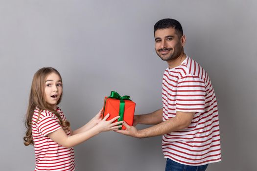 Portrait of delighted positive father and daughter in striped, celebrating little kid's birthday, dad giving present box to amazed child. T-shirts Indoor studio shot isolated on gray background.