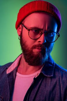 Closeup portrait of pensive hipster man in glasses, standing looking away, expressing thoughtfulness, wearing beanie hat and denim vest. Indoor studio shot isolated on colorful neon light background.