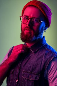 Portrait of attractive bearded hipster man in glasses, looking at camera, holding hand on his collar, wearing red beanie hat. Indoor studio shot isolated on colorful neon light background.