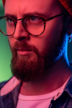 Closeup portrait of strict bossy bearded male in glasses looking to side space with serious attentive face, wearing beanie hat. Indoor studio shot isolated on colorful neon light background.
