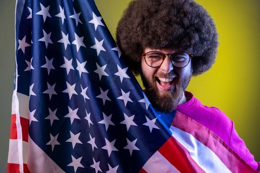 Portrait of hipster man with Afro hairstyle having fun, holding american flag, rejoicing, celebrating national holiday. Indoor studio shot isolated on colorful neon light background.