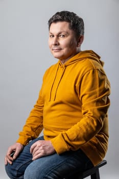 Portrait of attractive middle aged man looking at camera, sitting on chair and expressing positive emotions, wearing urban style hoodie. Indoor studio shot isolated on gray background.