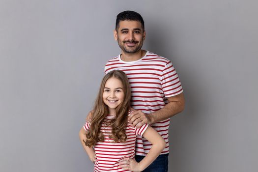 Portrait of happy positive father and daughter in striped T-shirts standing and looking smiling at camera, kid keeping hands on hips. Indoor studio shot isolated on gray background.
