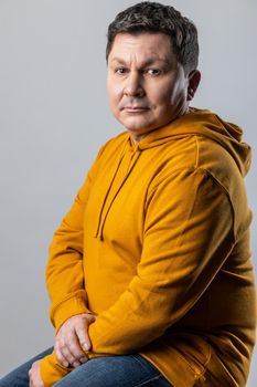 Portrait of confident handsome Caucasian man looking at camera, sitting on chair, looks calm, wearing urban style hoodie. Indoor studio shot isolated on gray background.