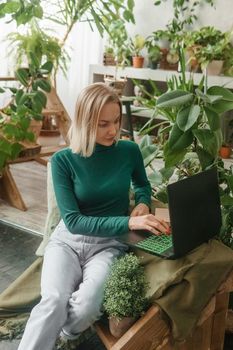 A blonde woman in a room with a lot of green indoor plants is working on a laptop. The concept of biophysical design in the interior. Work from home, work as a freelancer.