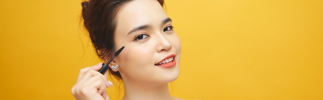 Closeup portrait of attractive young woman putting some mascara onto her eyelashes with make up brush over yellow background