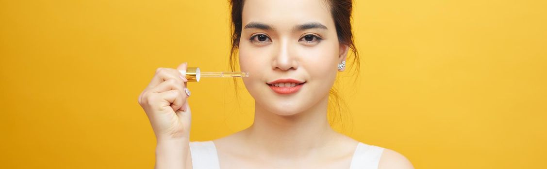 young beautiful asian woman hold pipette of serum for skin on yellow background