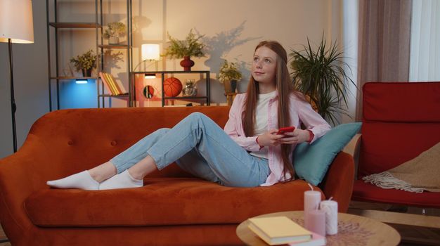 Young teen child girl lying on sofa uses mobile phone smile at modern home apartment. Ginger kid texting share messages content on smartphone social media applications online, watching relax movie