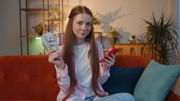 Planning family budget. Smiling young teen child girl showing money cash, use smartphone calculate domestic bills at home. Joyful kid satisfied of income and saves money for planned vacation, gifts