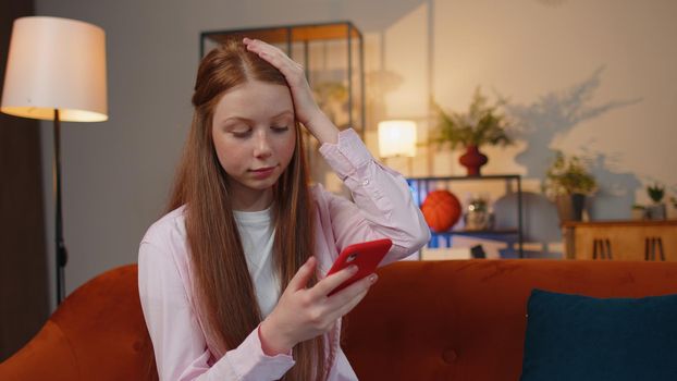 Young redhead teenager child girl use smartphone typing browsing, loses becoming surprised sudden lottery results, bad news, fortune loss, fail. Ginger female children kid at home living room couch