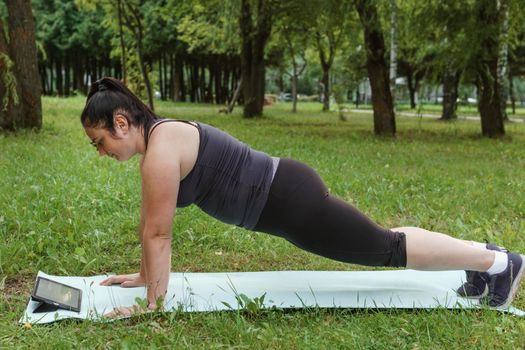 A charming brunette woman plus-size body positive practices sports in nature. Woman does yoga in the park on a sports mat. On a yoga mat in the dog pose.