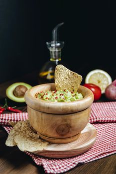 delicious guacamole in a wood bowl with tortilla chips next to avocado and other ingredients, typical mexican healthy vegan cuisine with rustic dark food photo style