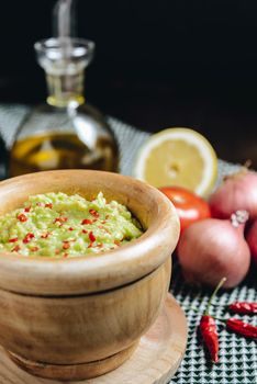 fresh guacamole in a wood bowl on a wooden table next to ingredients, typical mexican healthy vegan cuisine with rustic dark food photo style