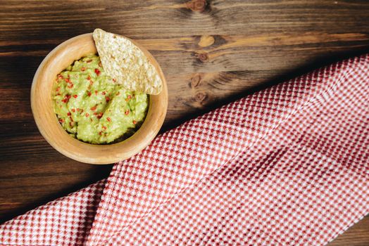 homemade guacamole with nachos in a wood bowl and a kitchen rag on a wooden table, typical mexican healthy vegan cuisine, top view and copy space for text