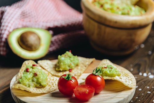 nachos with homemade guacamole in a wood plate next to an avocado and a kitchen rag, typical mexican healthy vegan cuisine with rustic dark food photo style, selective focus