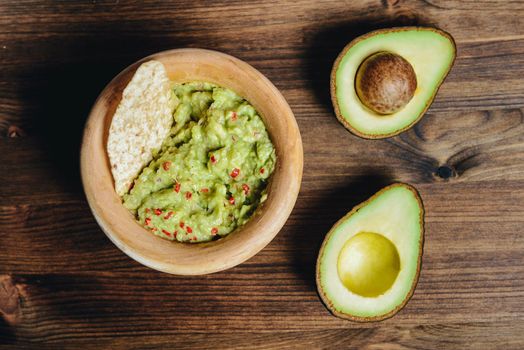 top view of traditional guacamole with nachos in a wood bowl and cut half avocado on wooden table, typical mexican healthy vegan cuisine with rustic dark food photo style