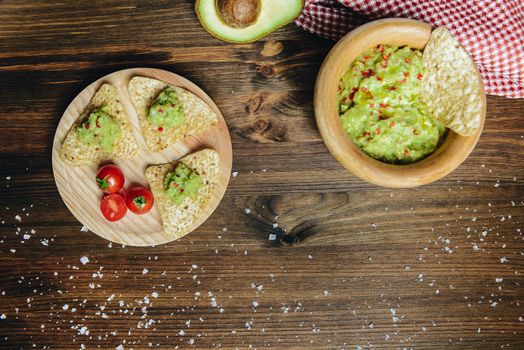 top view of homemade guacamole in a wood bowl with nachos next to an avocado and a kitchen rag, typical mexican healthy vegan cuisine with rustic dark food photo style