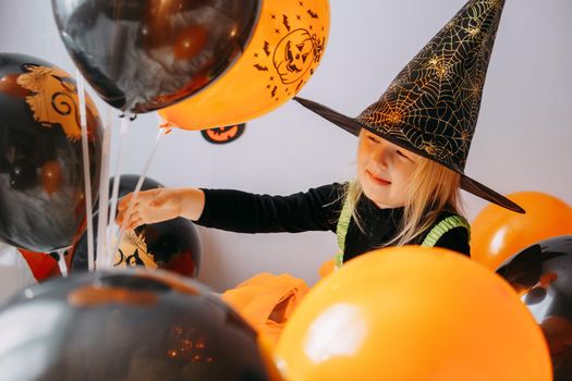 Children's Halloween - a girl in a witch hat and a carnival costume with airy orange and black balloons at home. Ready to celebrate Halloween.