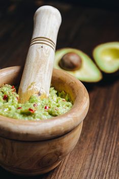 vertical photo of traditional guacamole in a wood bowl and cut half avocado on wooden table, typical mexican healthy vegan cuisine with rustic dark food photo style, selective focus