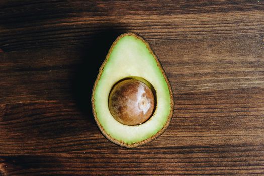 top view of half avocado with seed on wooden table, healthy vegan cuisine concept with rustic dark food photo style