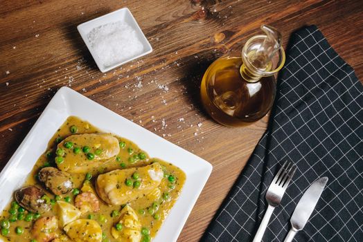 healthy traditional spanish hake dish with green seafood sauce with clams, prawns, green peas and egg, top view of typical mediterranean cuisine with rustic dark food style