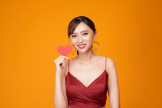happy young woman dressed in red dress holding paper heart isolated over yellow background
